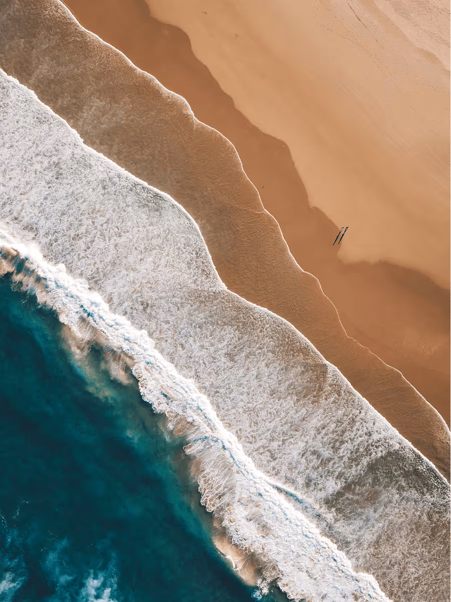Aerial view of ocean waves crashing onto a sandy beach with two small figures standing on the shore.
