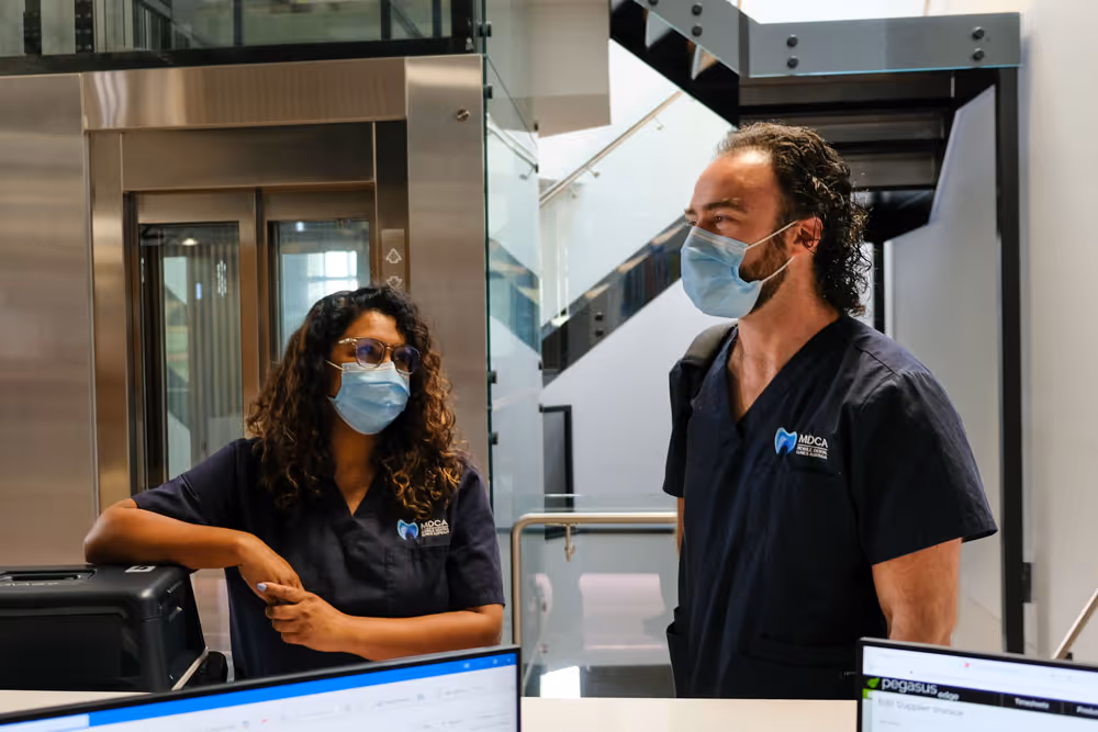 Two masked dental professionals in navy scrubs talking at a clinic reception with computers and elevator.