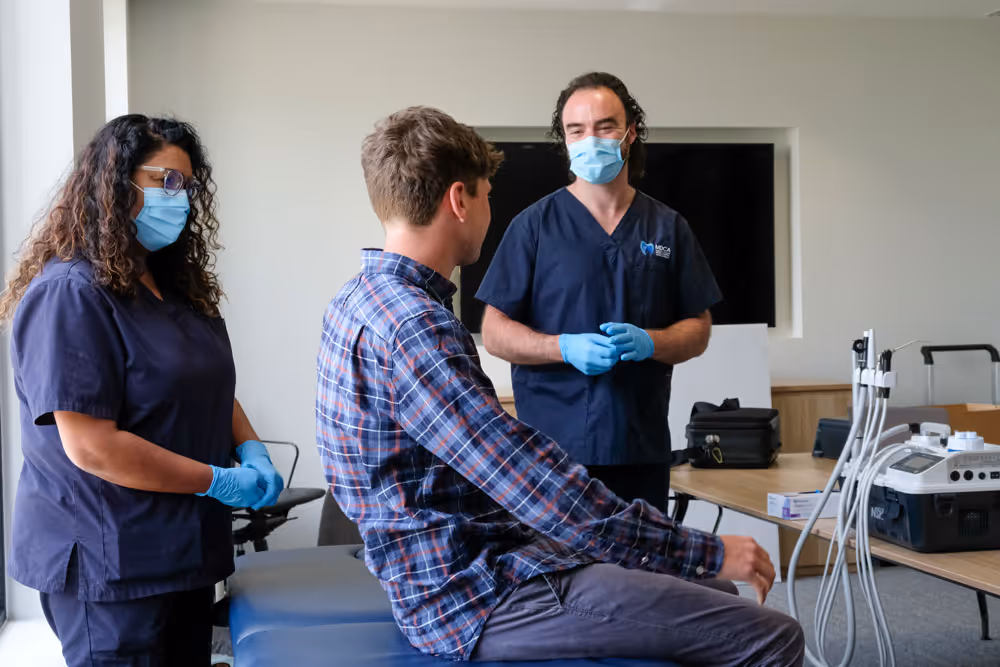 Two healthcare workers in scrubs and masks talk with a male patient sitting on an exam table.