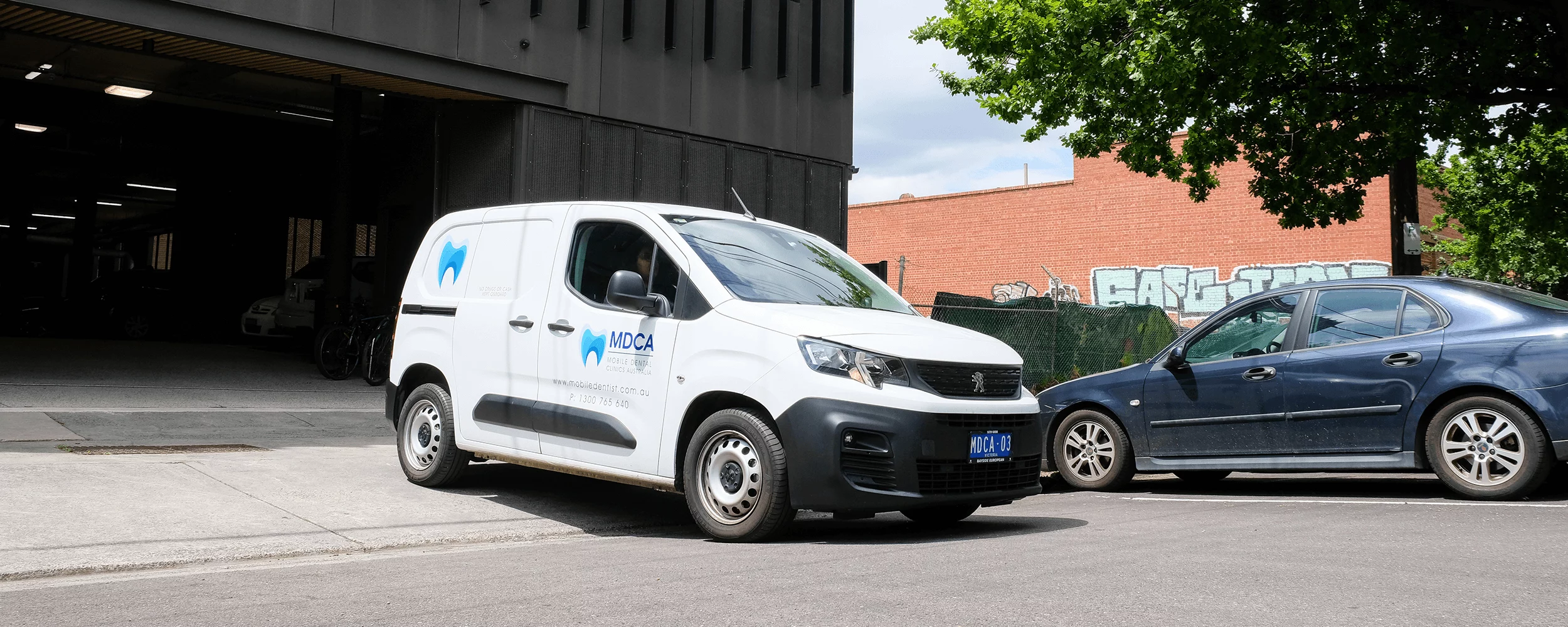 White MDCA Mobile Dental Clinics Australia van parked near a blue sedan in an urban setting.