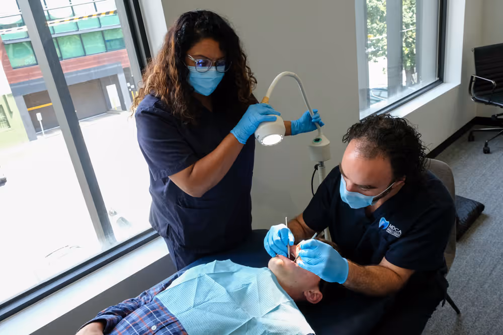 Dentists wearing masks and gloves treat patient lying on dental chair near window.
