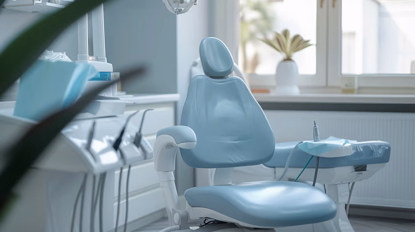 Empty blue dental chair and equipment in a well-lit dental office room with windows.