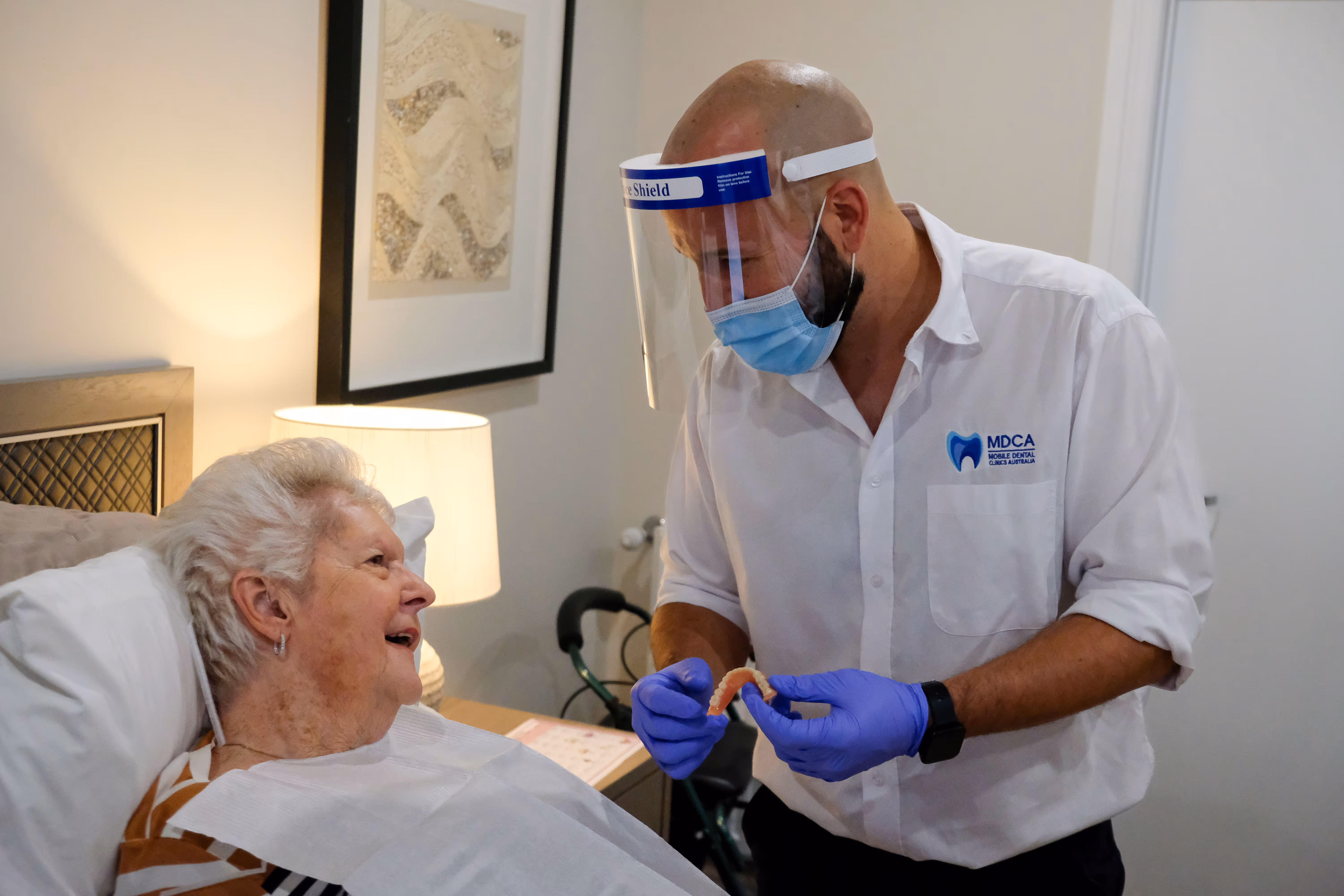 Dentist wearing face shield and mask showing dentures to elderly woman in bed.