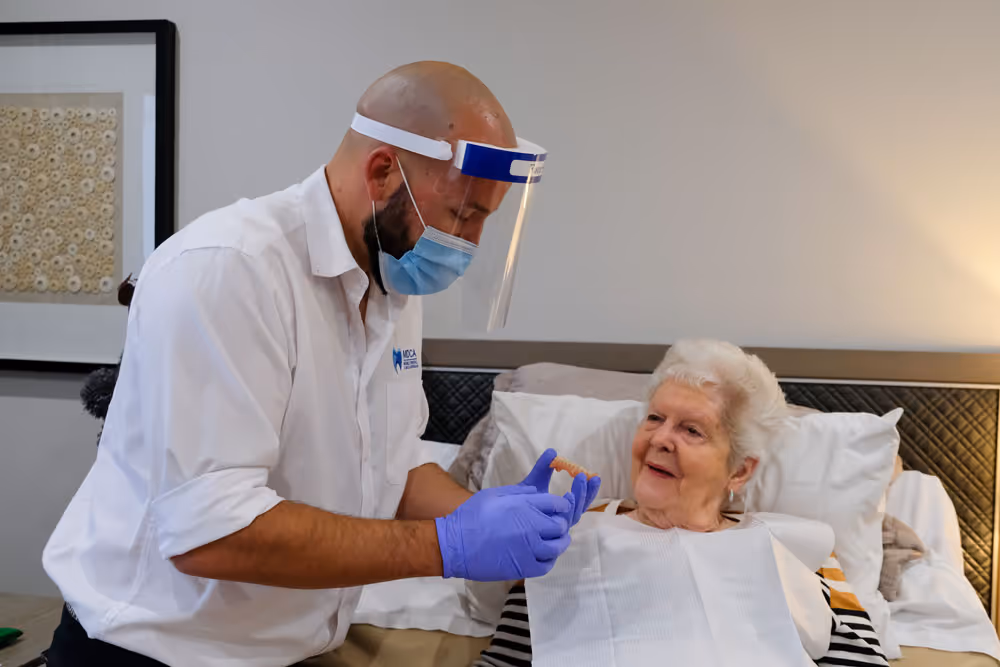 Healthcare worker in mask and face shield assists elderly woman with dentures in a home care setting.