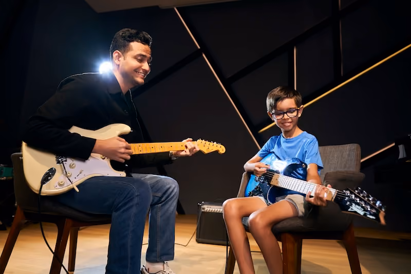 A music teacher holding an electric guitar is seated across from his student who is also holding an electric guitar. They are both smiling. 
