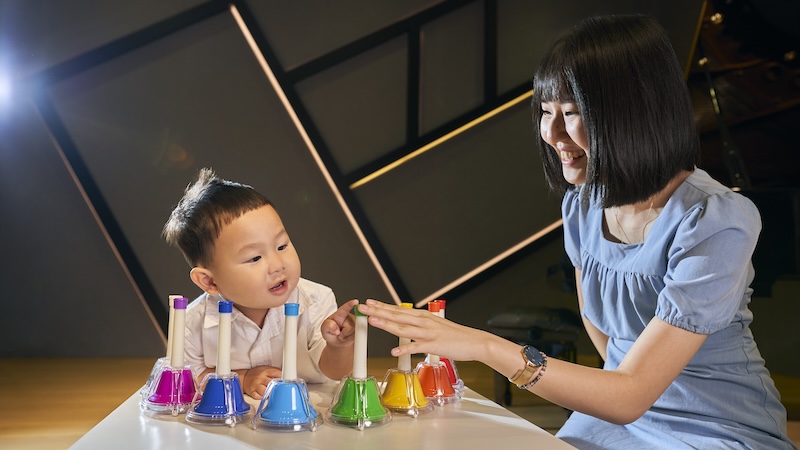 Music exploration class with a young kid and teacher, in blue. Both are reaching out to hit a green bell.
