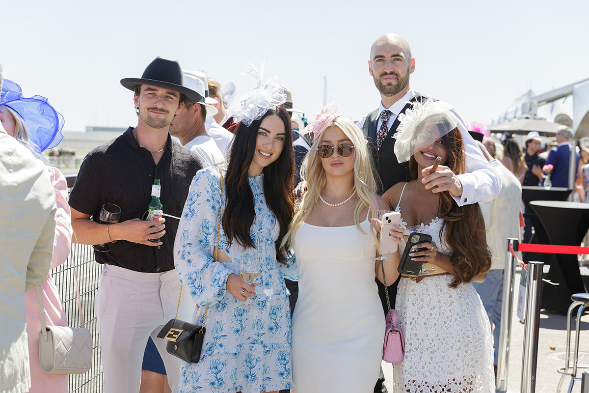 Group of five stylishly dressed people at an outdoor event, three women wearing elegant dresses and fascinators, two men in smart casual attire, one holding a cigar.