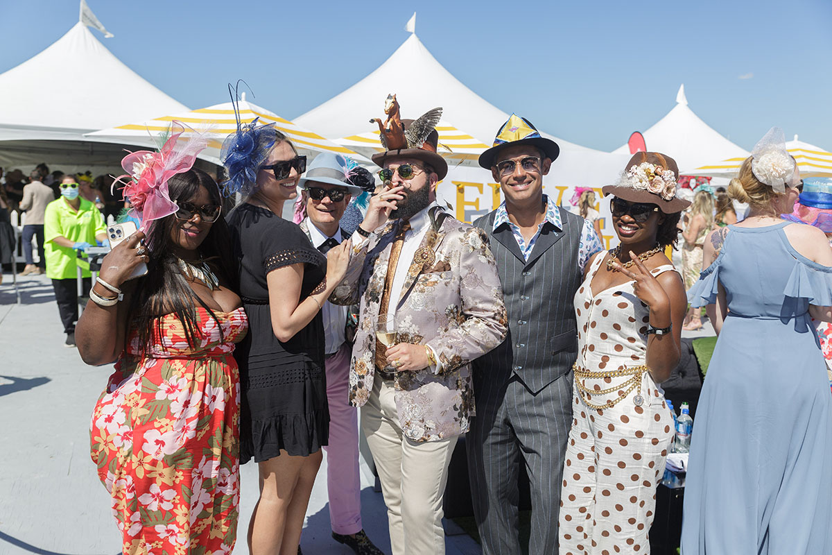 Group of six people dressed in colorful, stylish outfits and hats posing outdoors at a daytime event with white tents and blue sky in the background.