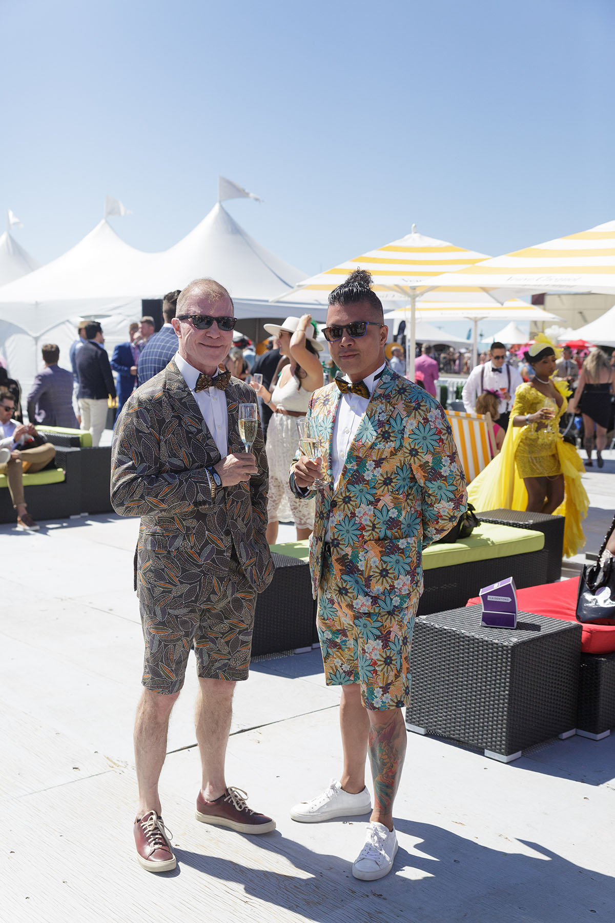 Two men wearing colorful patterned short suits and sunglasses holding champagne glasses at an outdoor event with white tents and umbrellas.