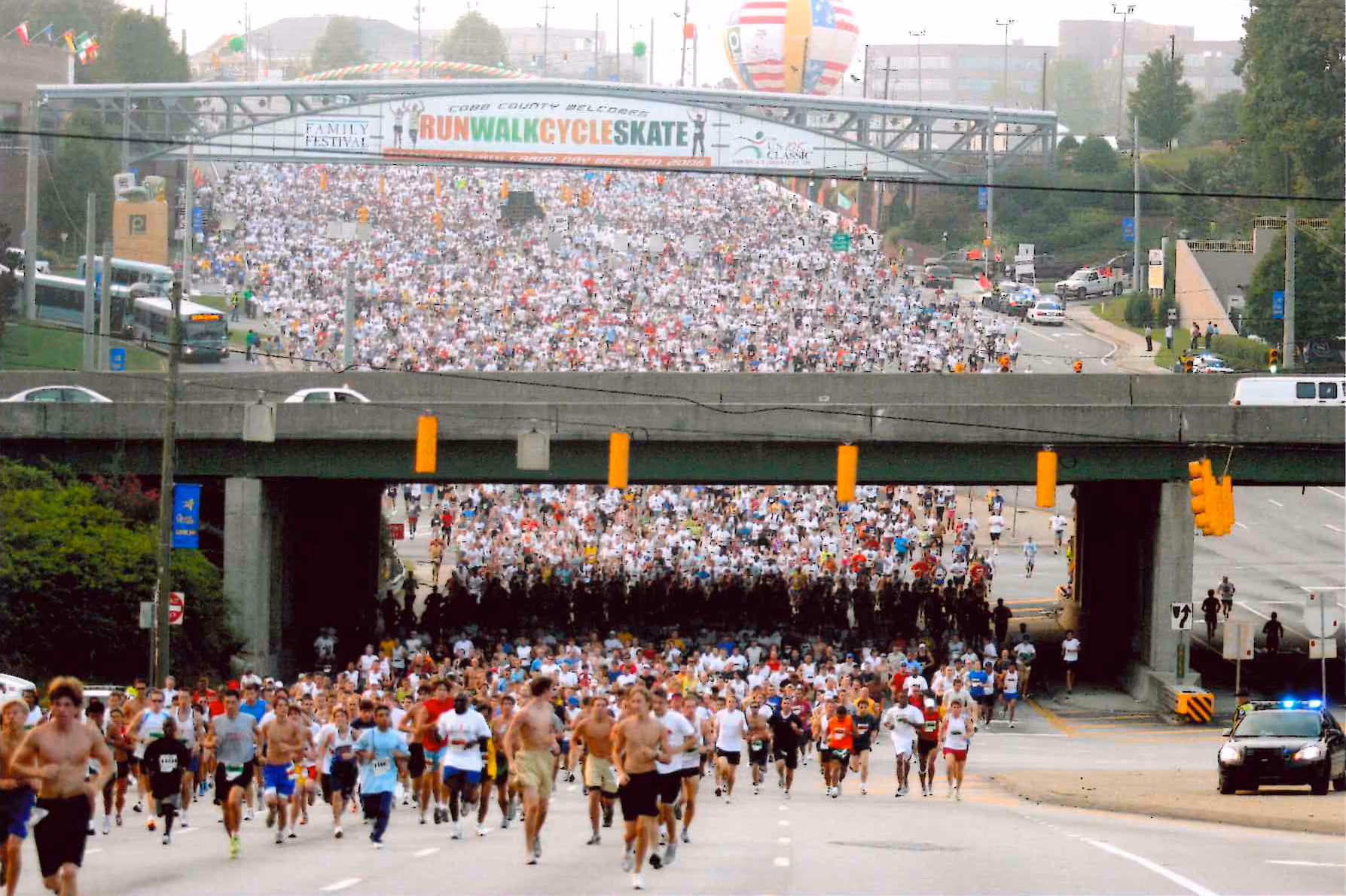 Large crowd of runners participating in a race under an overpass with a banner reading 'Run Walk Cycle Skate' in the background.