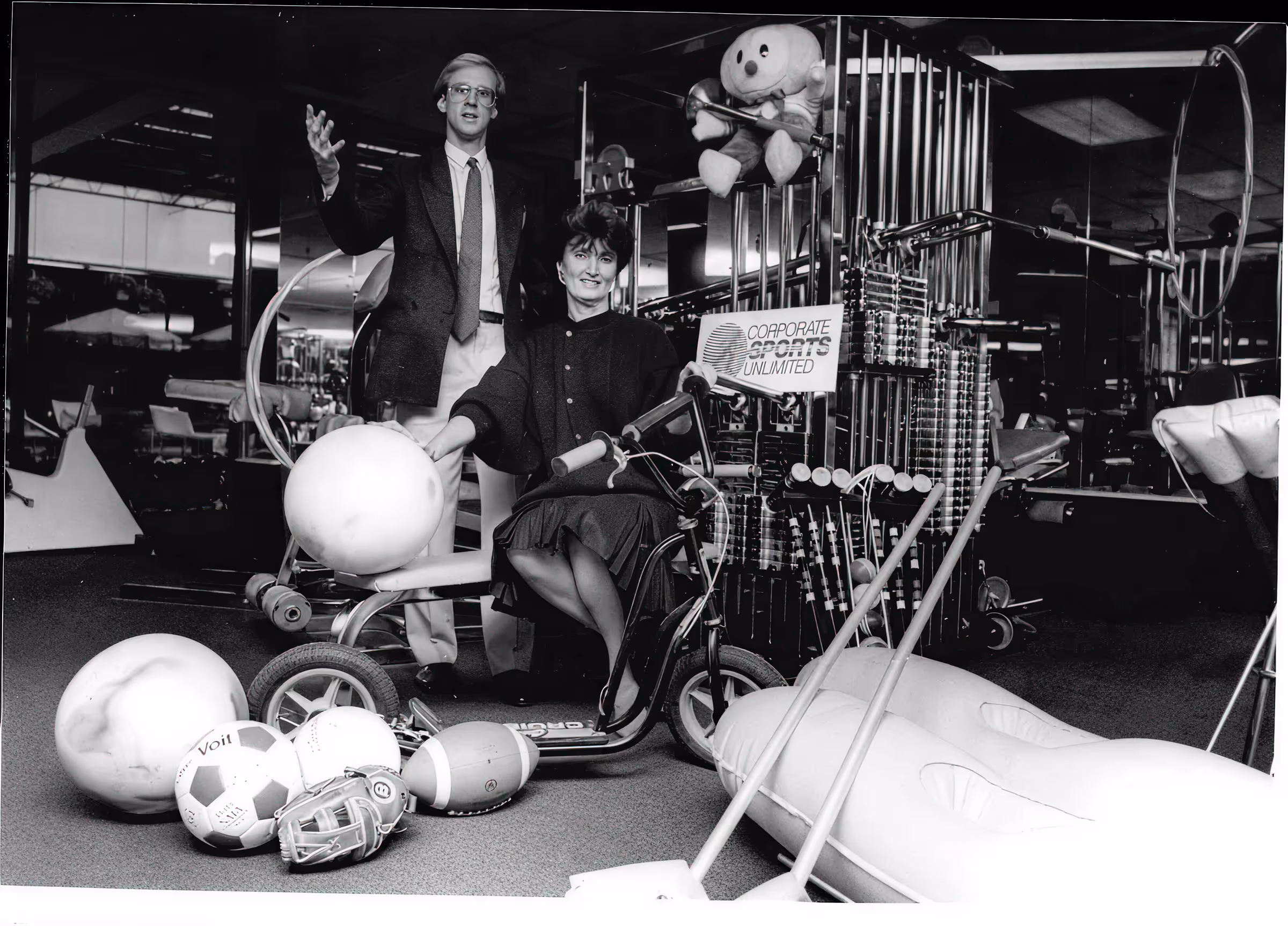 Man and woman in a gym surrounded by various sports balls, exercise equipment, and a scooter with a sign reading Corporate Sports Unlimited.