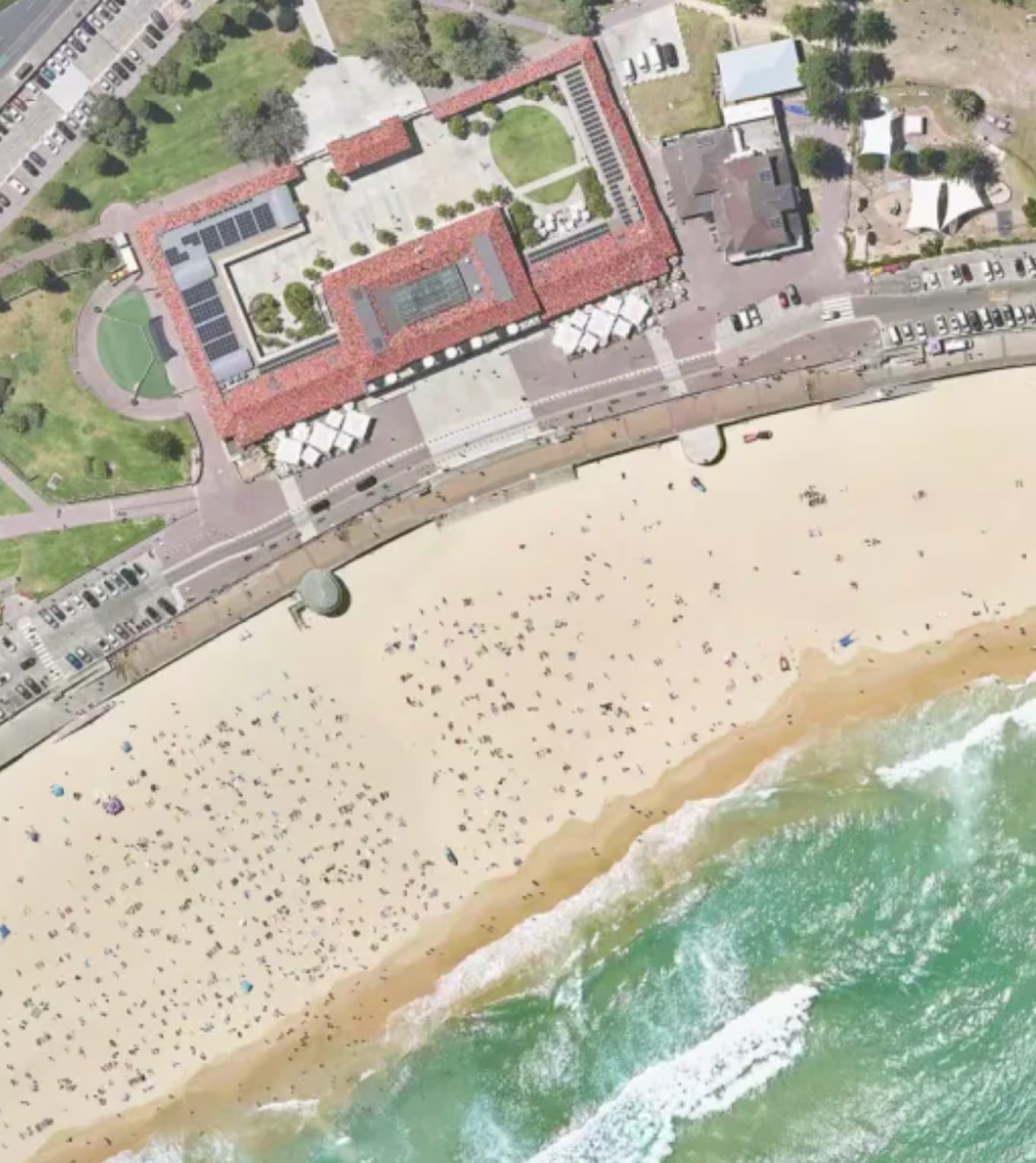 Aerial view of a sandy beach with scattered people, ocean waves, and adjacent buildings and parking lots.