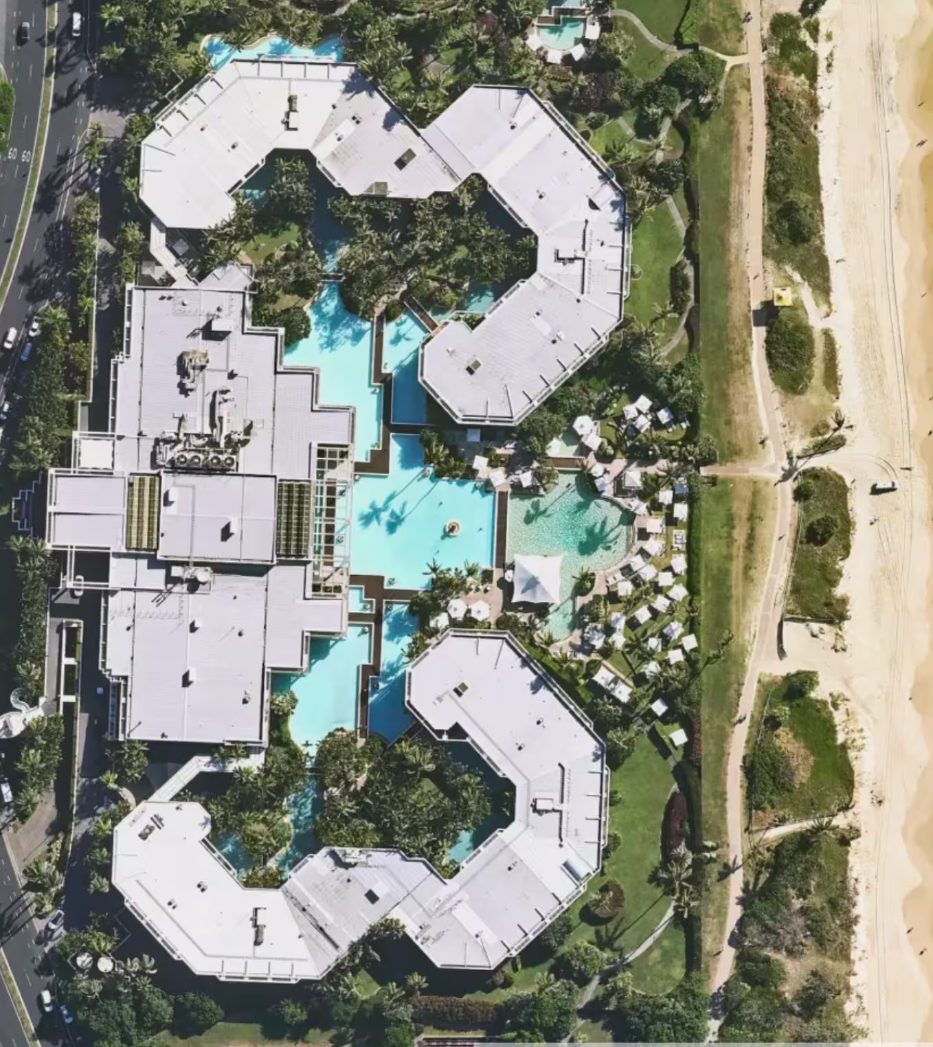 Aerial view of a resort with pools, white-roofed buildings, palm trees, and beach on the right side.