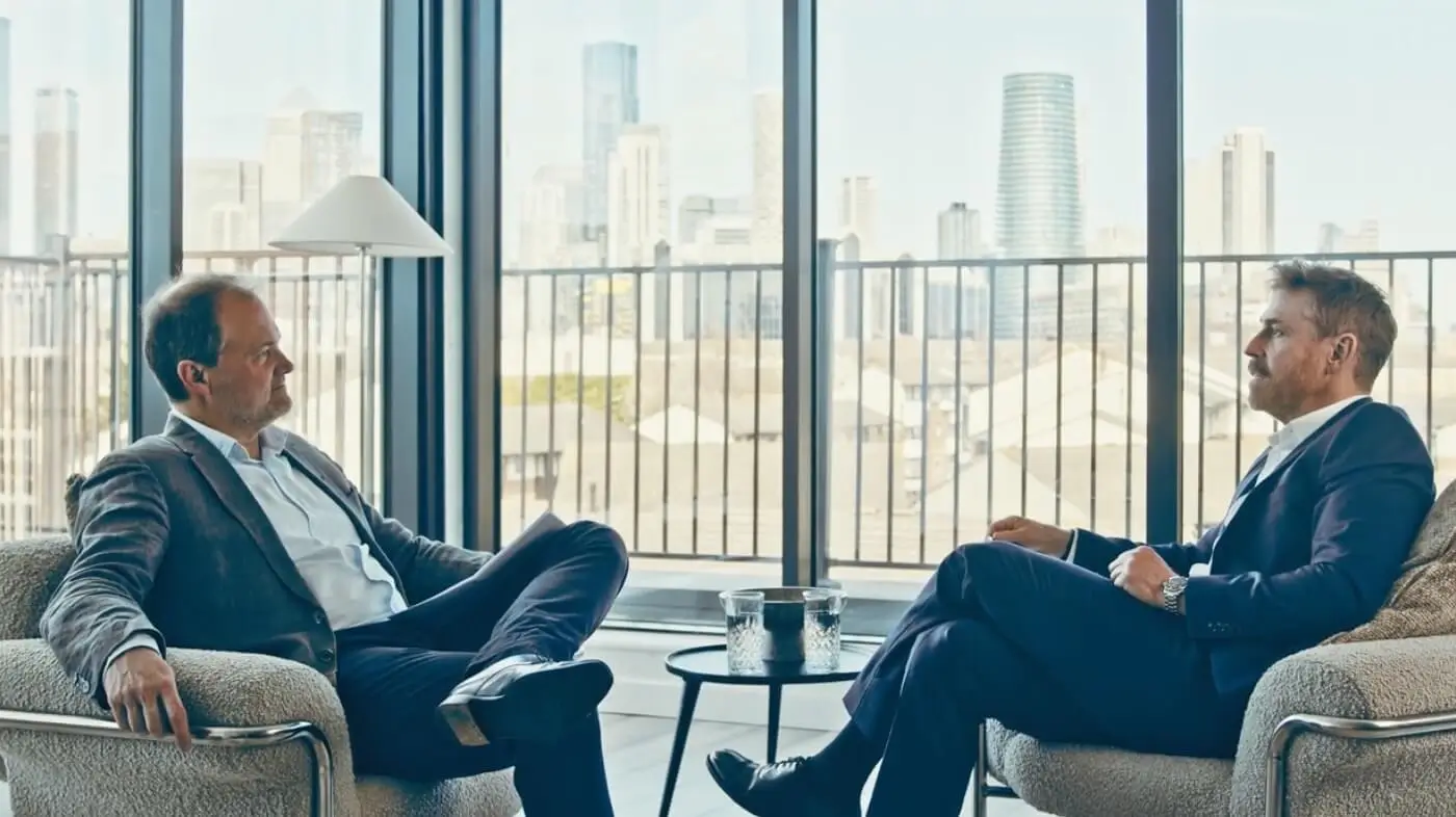 Two men in suits sitting on armchairs facing each other with a city skyline visible through large windows.