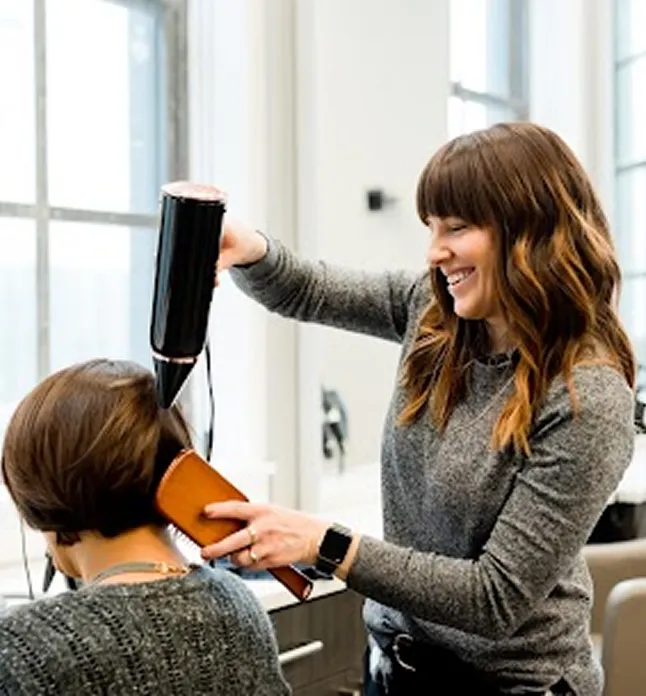 Smiling hair stylist blow-drying a client's short brown hair in a bright salon.