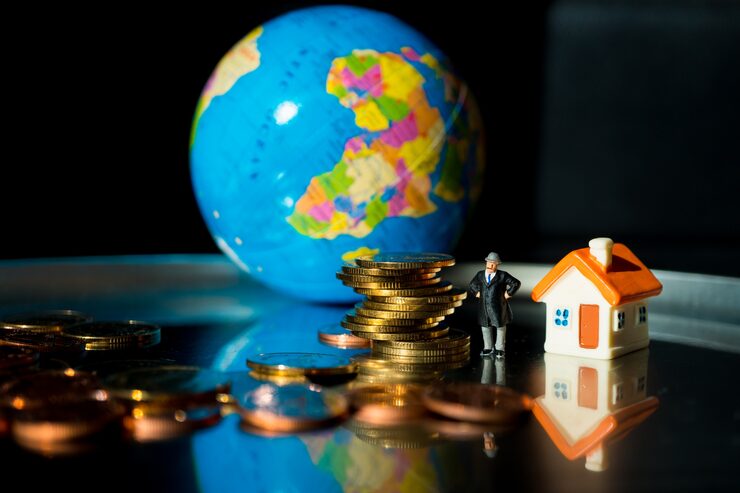 Miniature man stands beside coins and a small house figurine with a globe in the background.