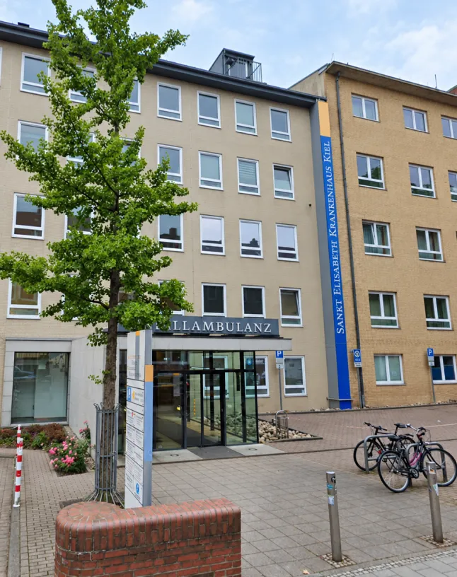 Lächelnder Arzt mit Brille, weißem Kittel, roter Krawatte und Stethoskop hält ein Klemmbrett.