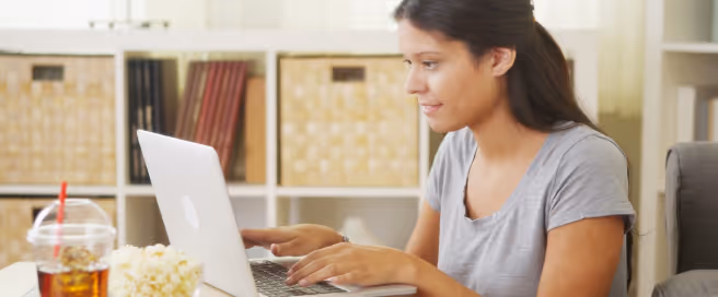 Young woman with dark hair tied back, wearing a light gray t-shirt, typing on a silver laptop in a cozy room with shelves and storage boxes in the background.