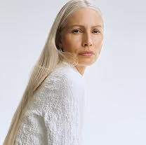 Portrait of an older woman with long gray hair wearing a white textured sweater, looking thoughtfully at the camera against a plain light background.