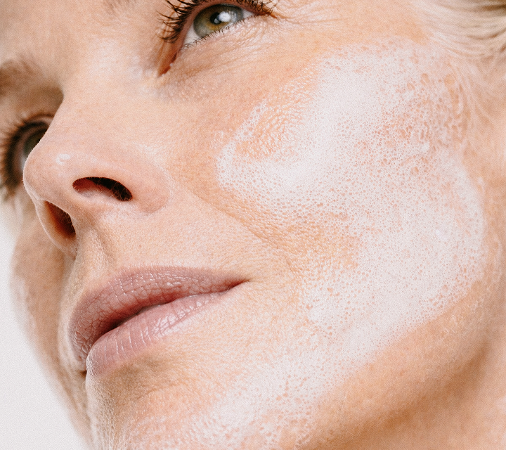 Close-up of a woman's face with clear skin, showing a foamy white facial cleanser applied to her cheek, looking slightly upwards.