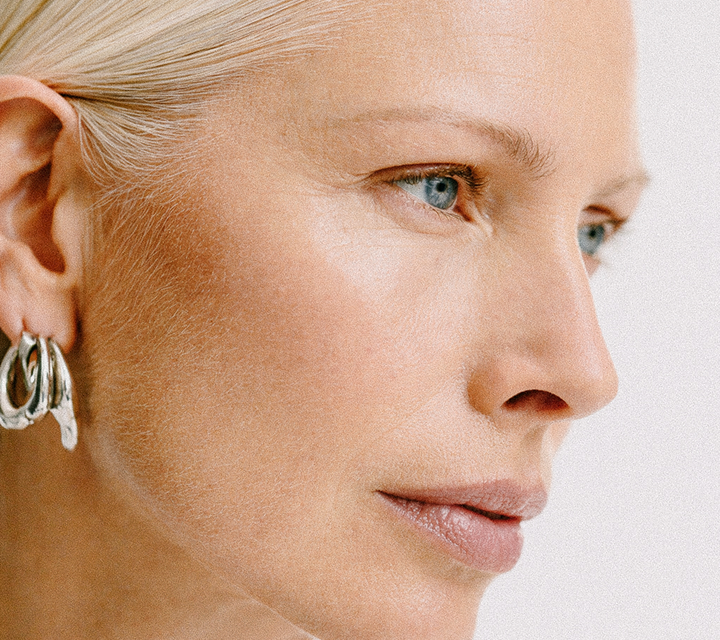 Close-up profile of a woman with fair skin and blue eyes looking to the right, wearing silver hoop earrings and with blonde hair pulled back.