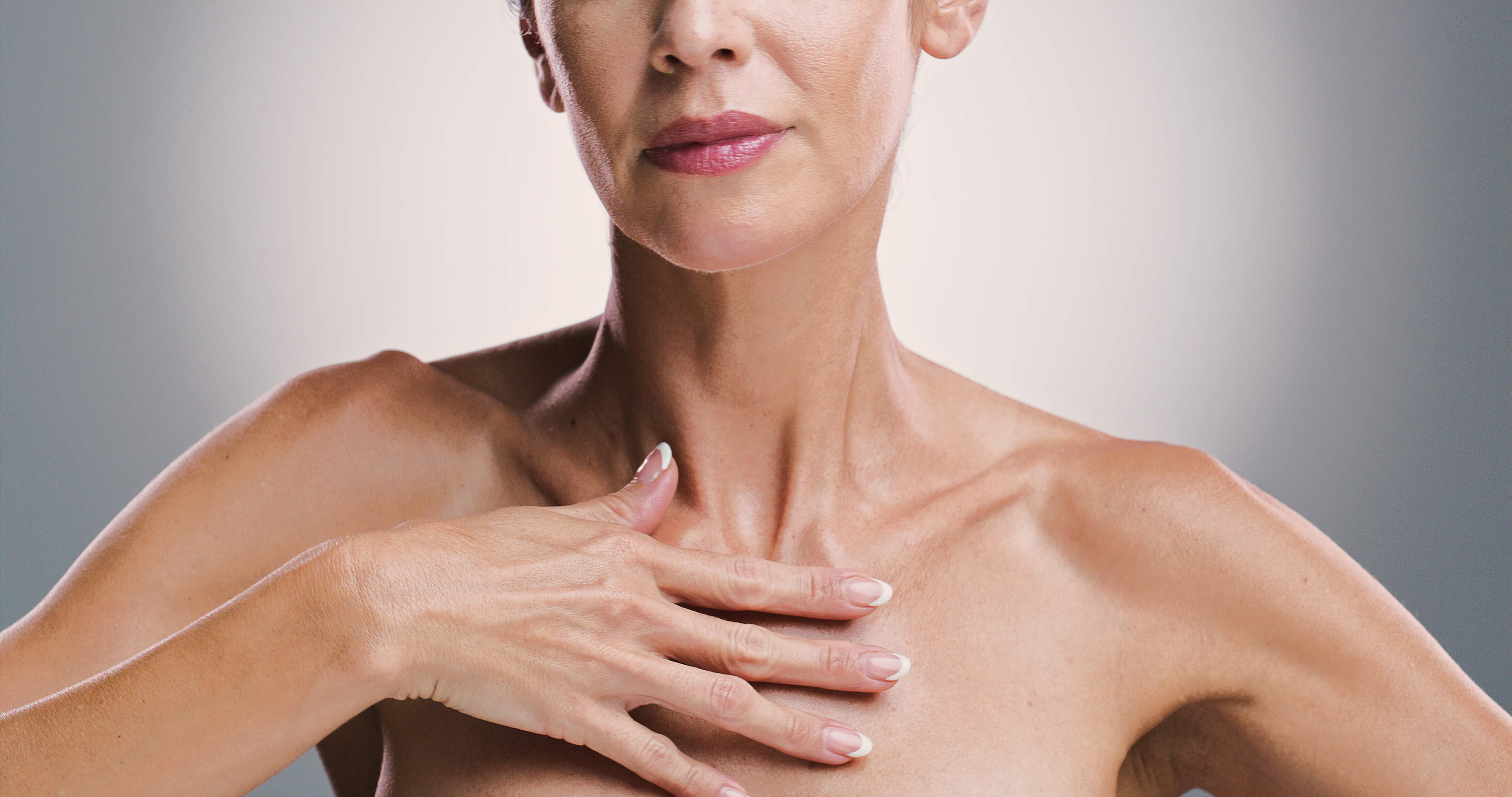 Close-up of a woman’s neck, upper chest, and lips with her hand gently touching her chest, showing smooth skin and a well-manicured French manicure.