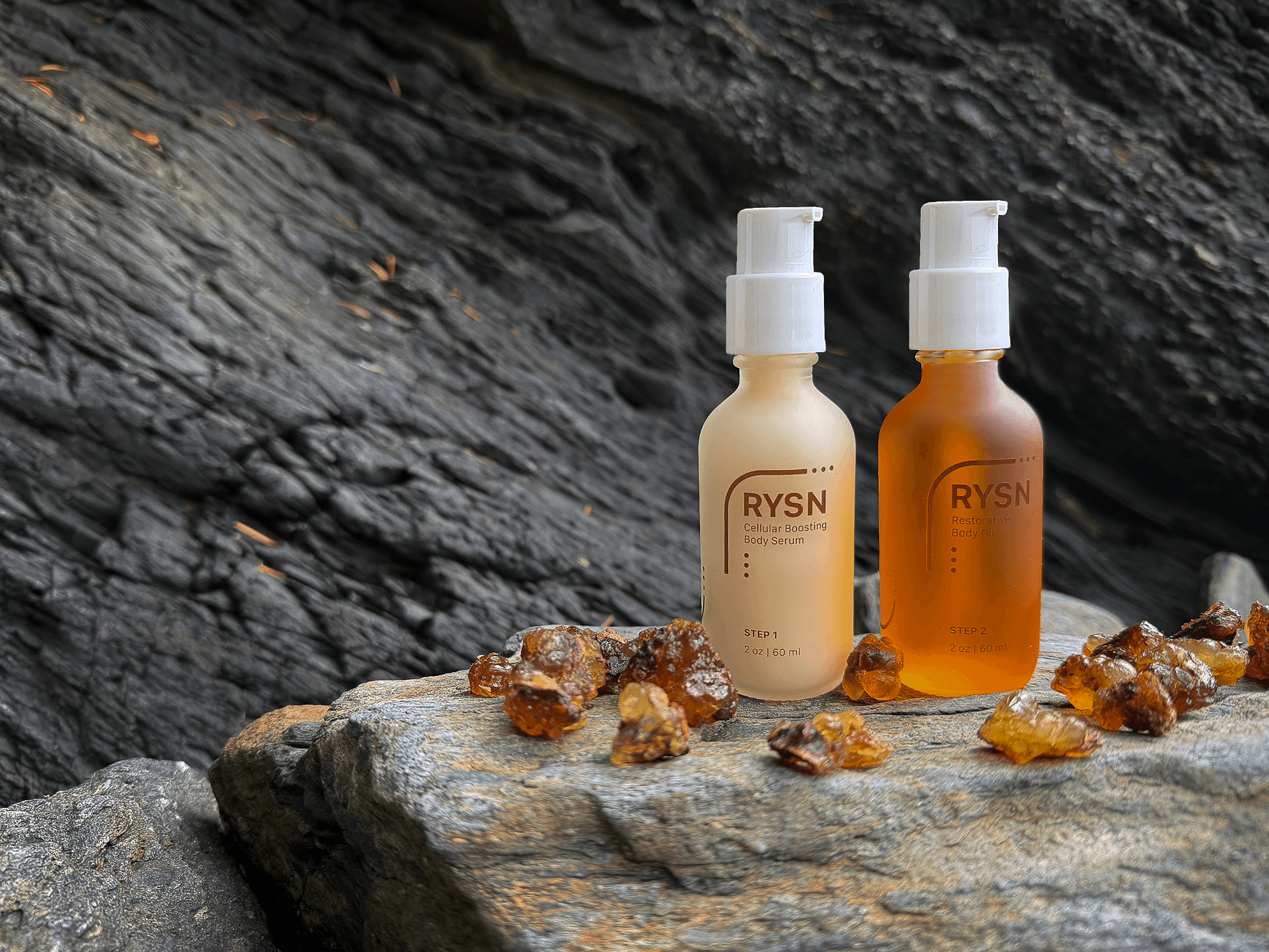 Two RYSN branded skincare bottles, one labeled Cellular Boosting Body Serum and the other Restorative Body Oil, placed on a rocky surface with amber resin pieces scattered around them against a dark textured rock background.