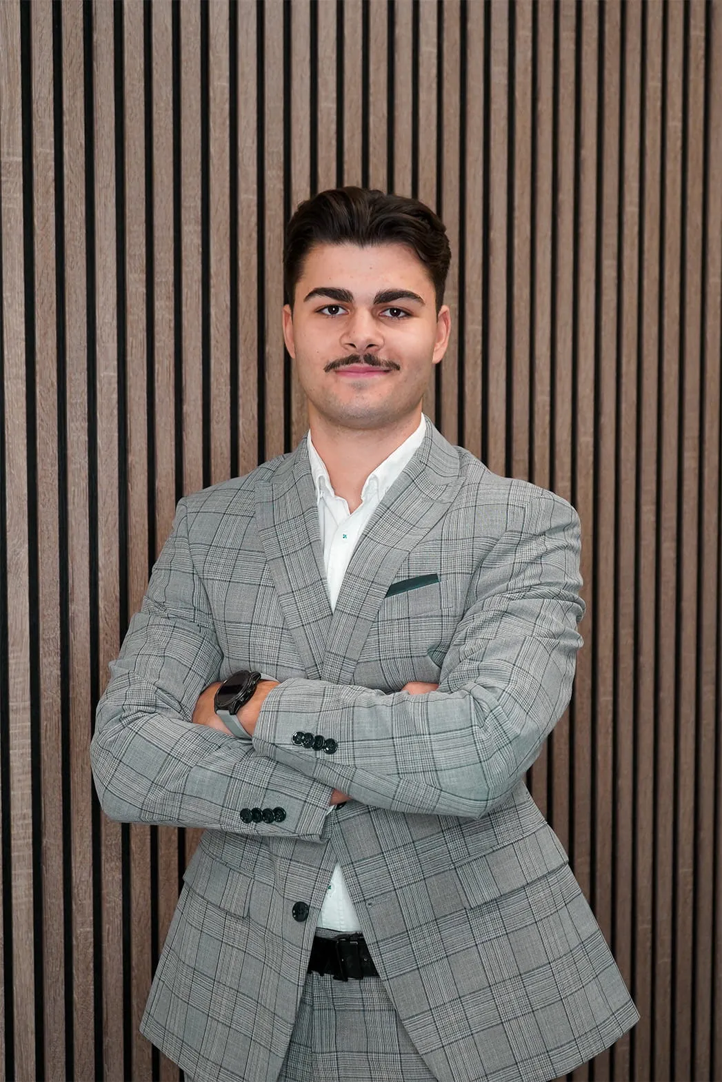Young man with a mustache wearing a grey plaid suit with arms crossed standing in front of a vertical wooden slat wall.
