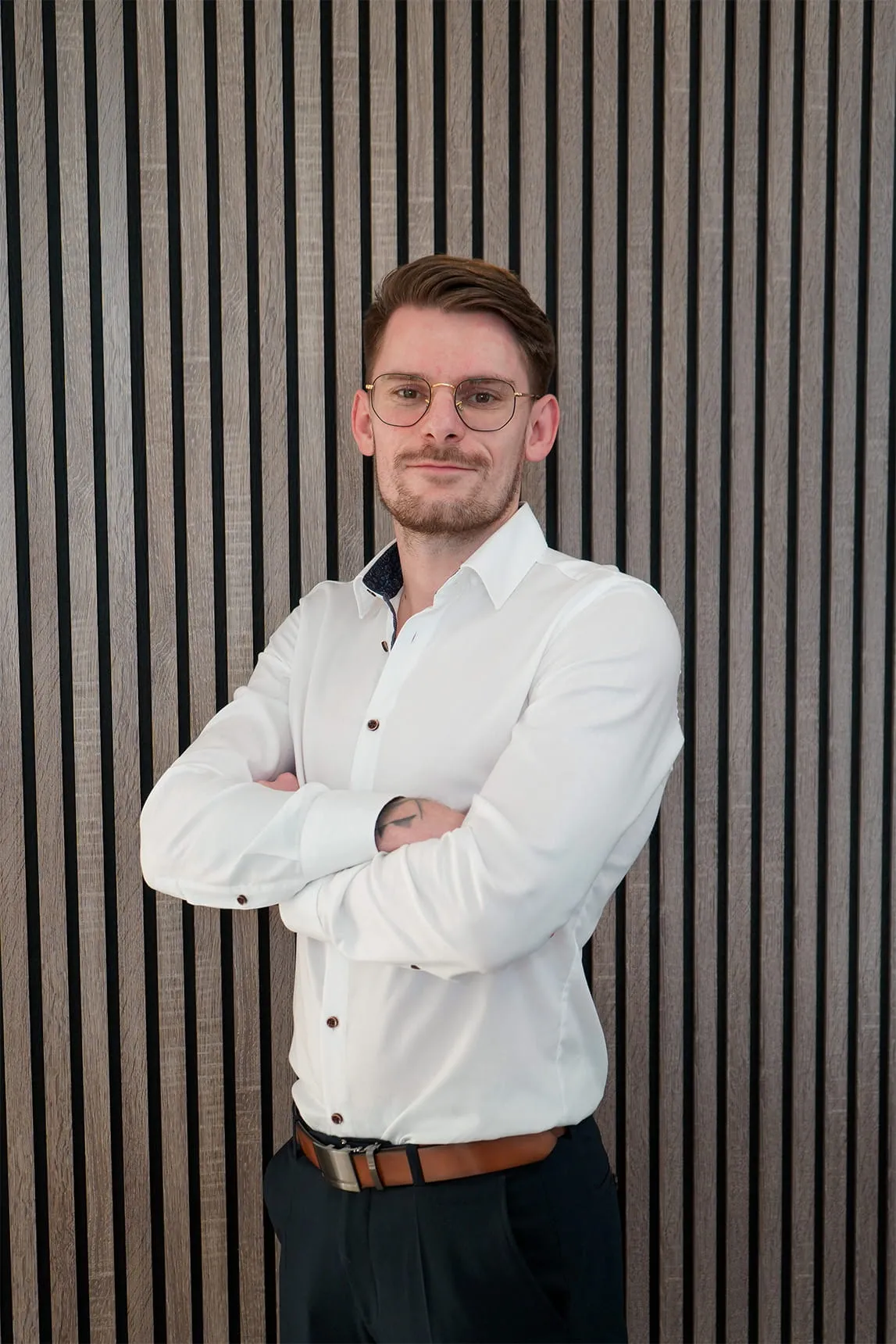 Young man with glasses, wearing a white shirt and black pants, standing with arms crossed in front of a vertical striped wooden wall.