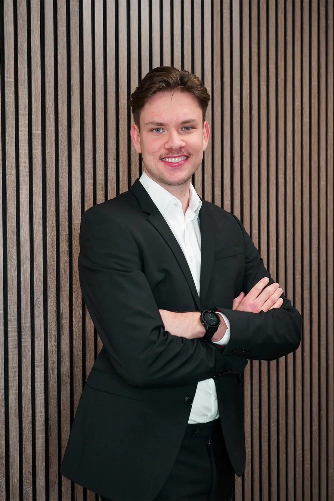 Young man in a black suit and white shirt smiling with arms crossed against a vertical wooden slat wall.