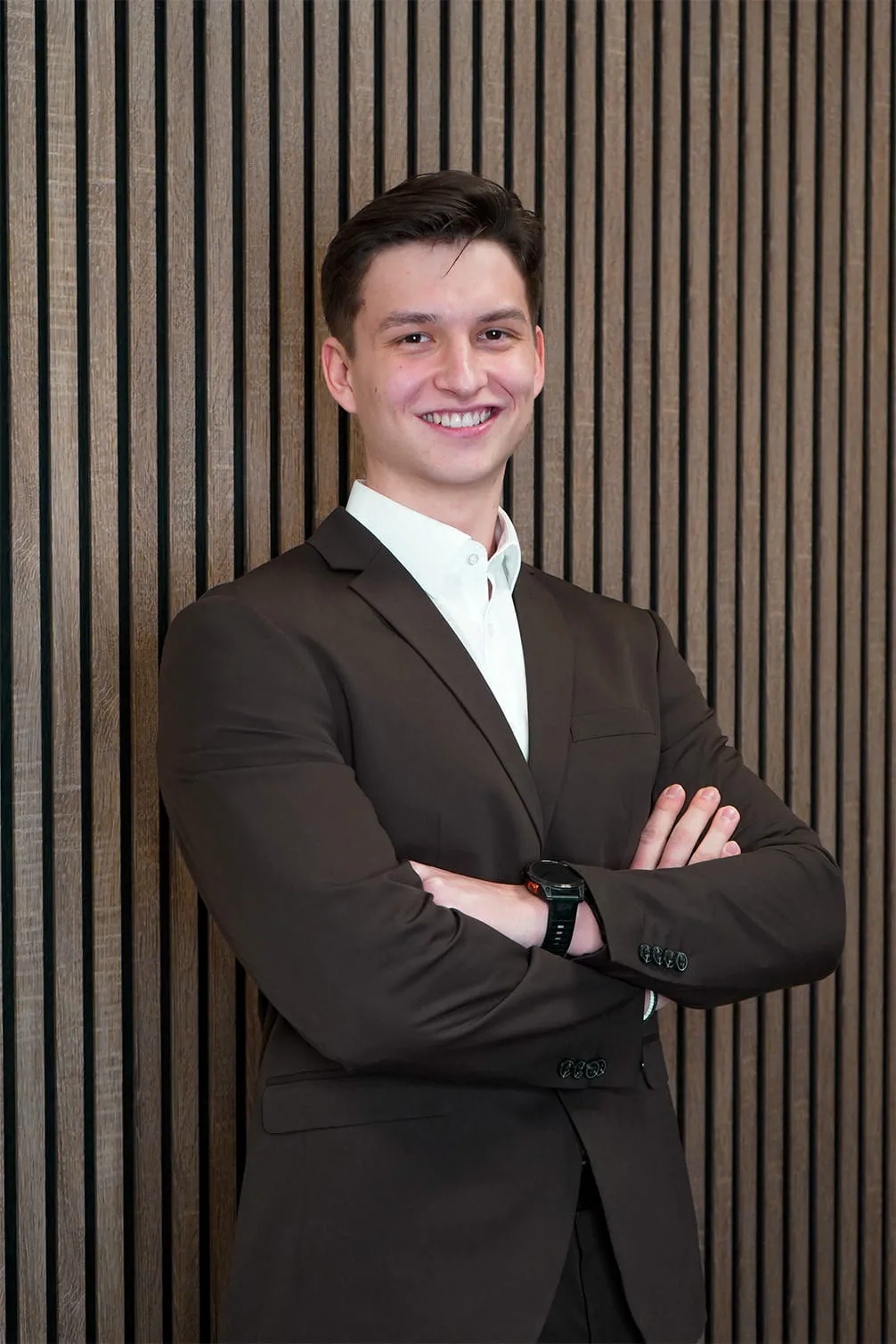 Smiling young man in a dark suit with arms crossed standing in front of a vertical wooden panel wall.