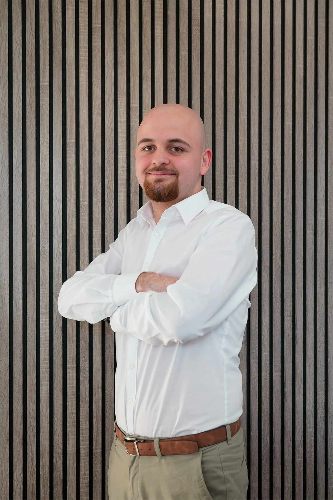 Smiling bald man with a goatee wearing a white dress shirt and beige pants, standing with arms crossed in front of a wood panel wall.