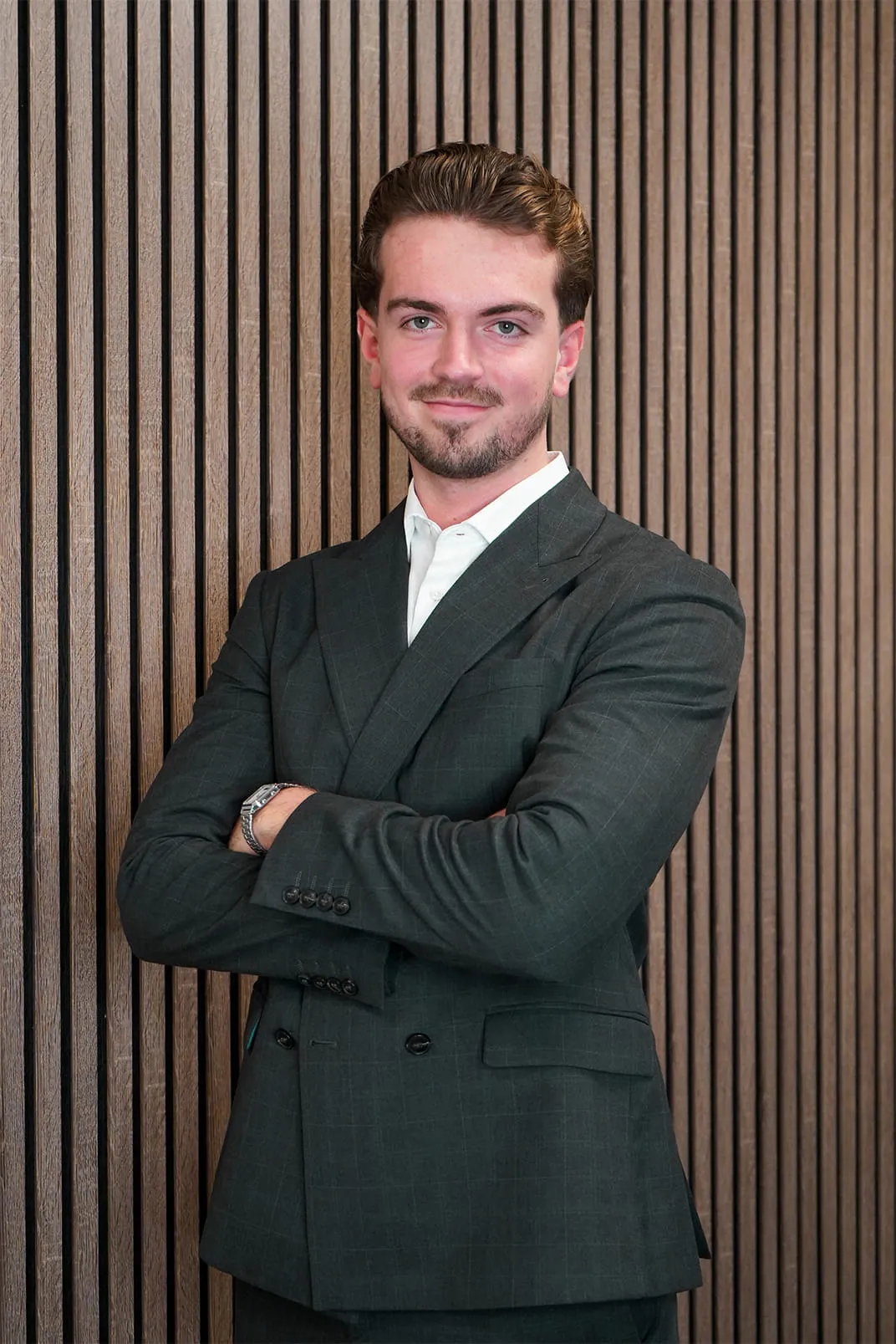 Smiling young man with neatly combed hair wearing a dark suit and white shirt, standing with arms crossed in front of a vertical wooden panel wall.