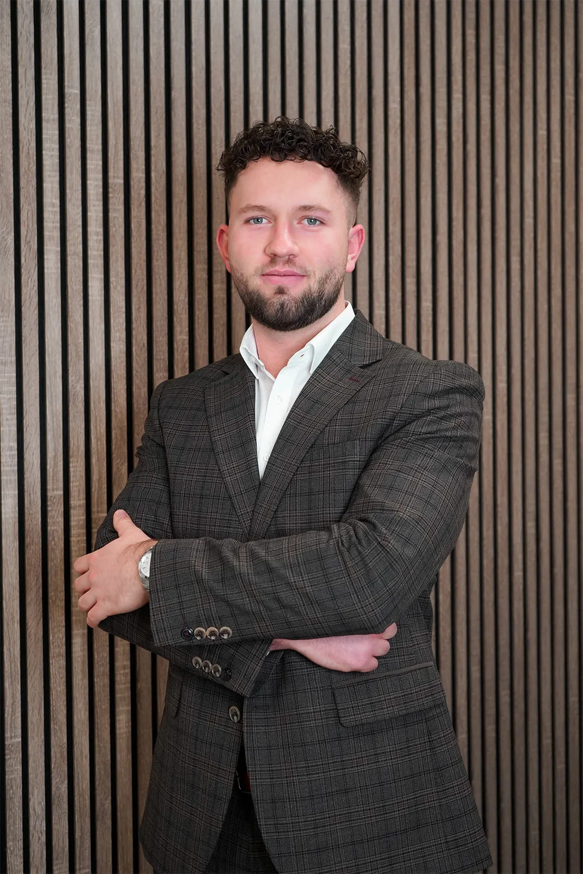 Man with curly hair and beard wearing a plaid suit jacket and white shirt standing with arms crossed in front of vertical wooden slats.