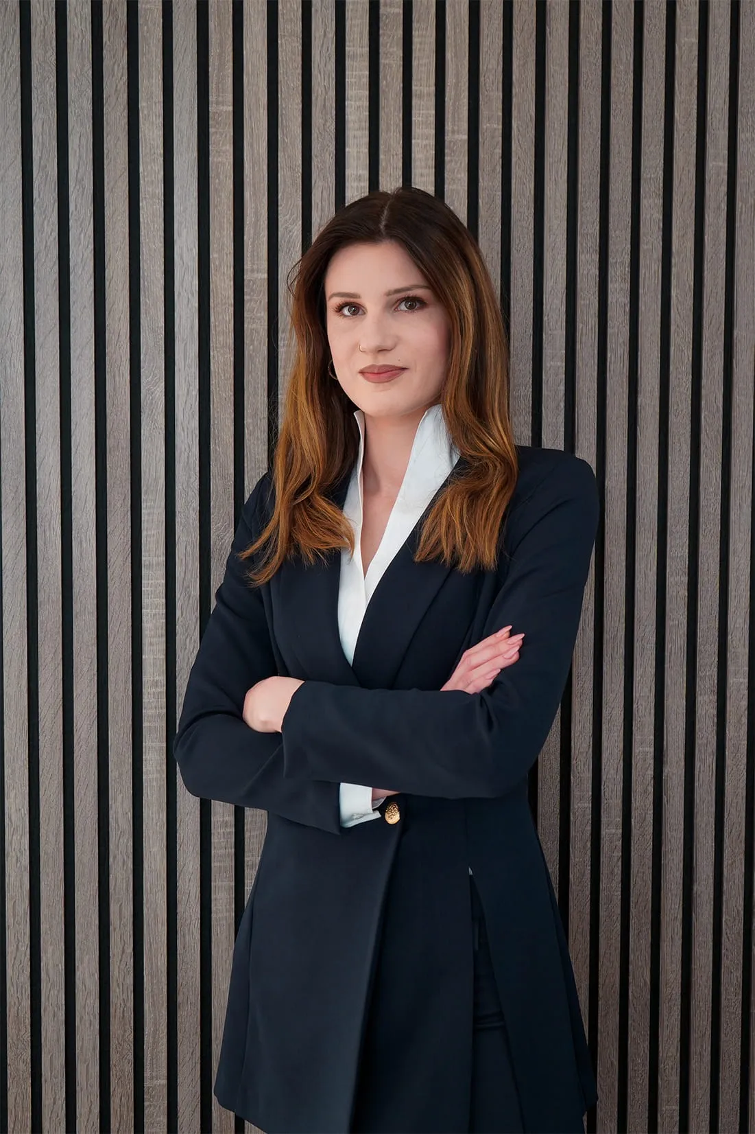 Confident woman with crossed arms wearing a navy blazer and white shirt standing against a striped wood panel background.