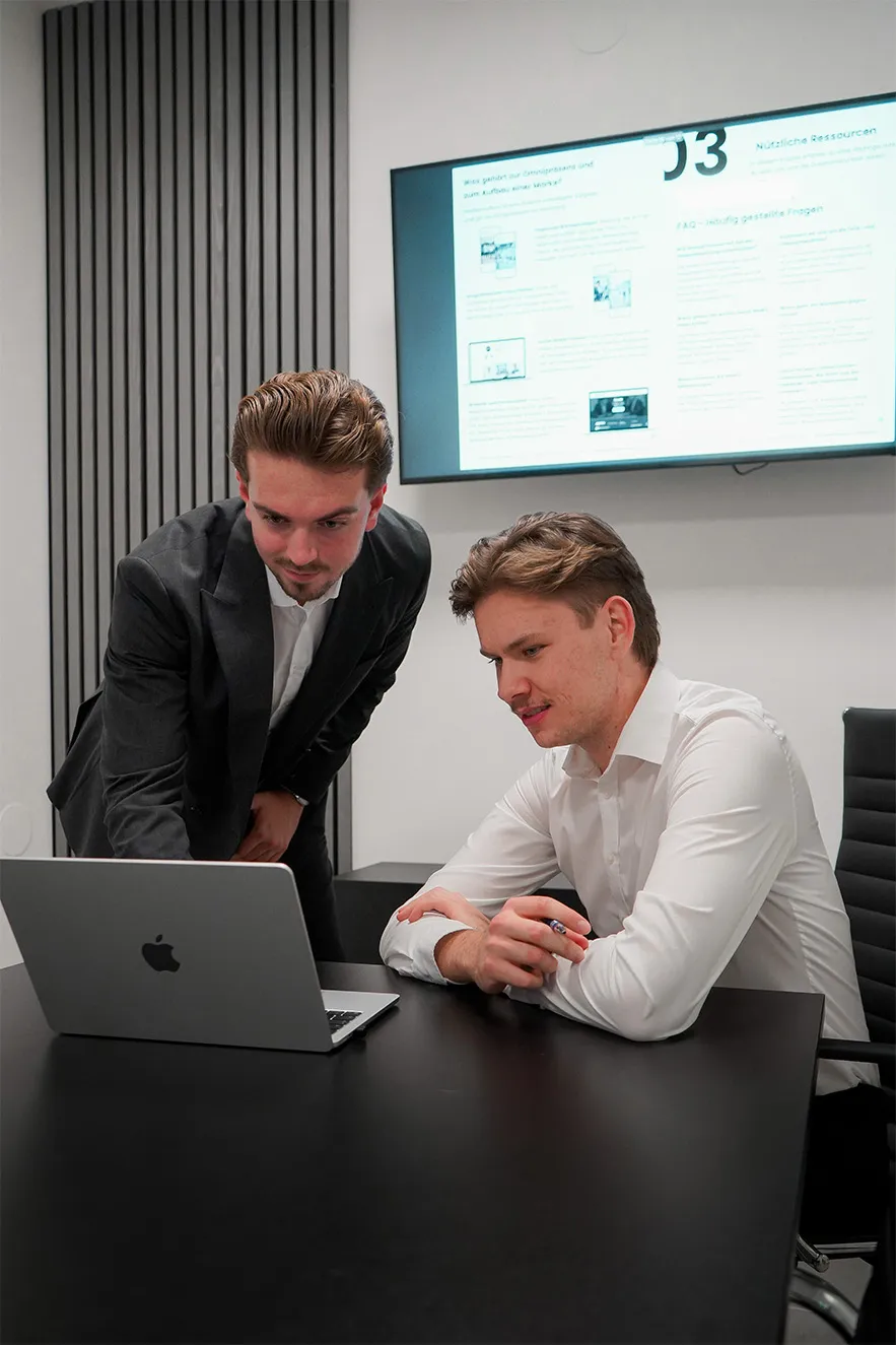 Two men in business attire looking at a laptop in a modern office with a screen on the wall.