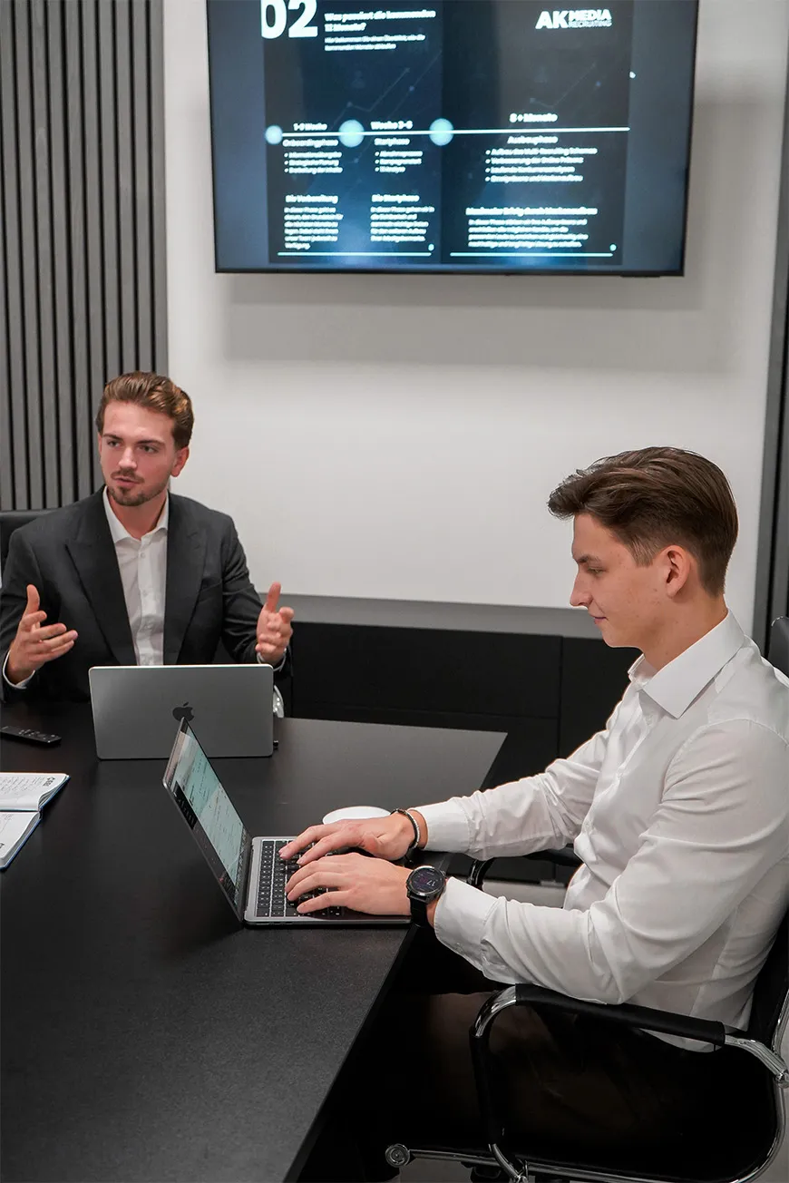 Two men in a meeting room with laptops, one talking and the other typing, screen with presentation in background.