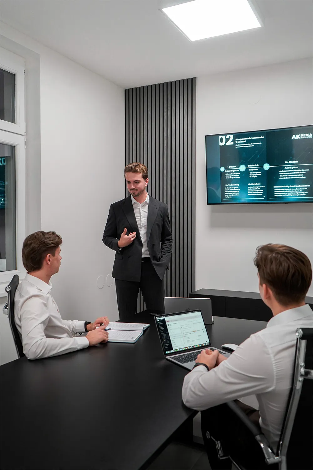 Man in suit stands explaining to two seated men during a meeting in a modern office with a wall screen.
