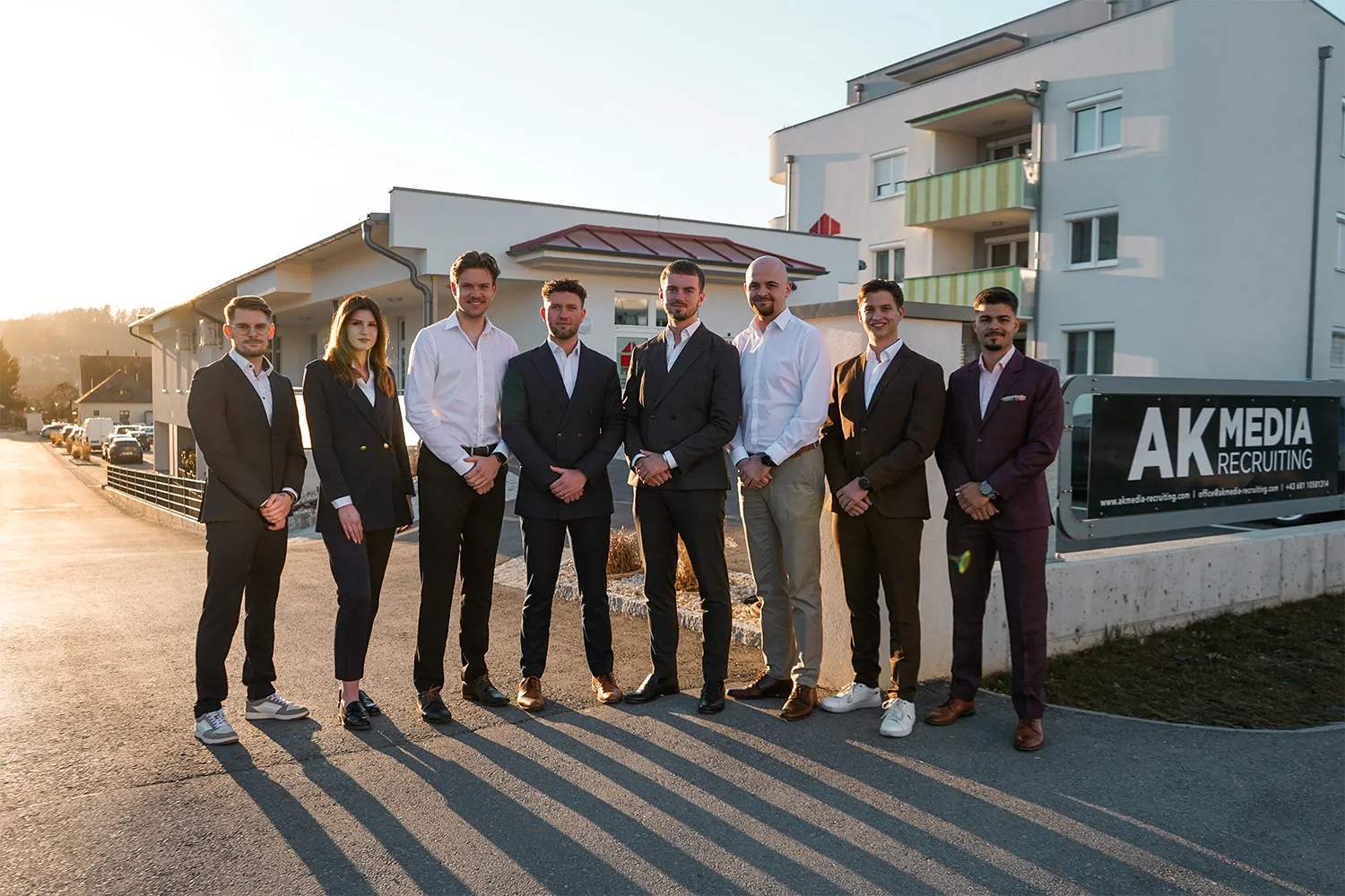 Eight professionals in suits standing outside an office building with AK Media Recruiting sign.