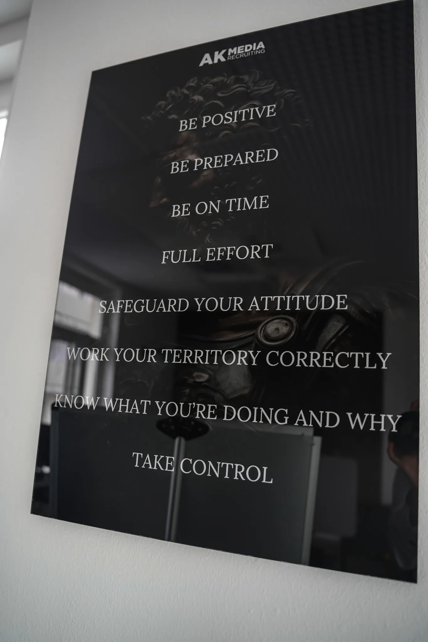 Black wall sign with AK Media logo listing work principles like be positive, prepared, on time, and take control.