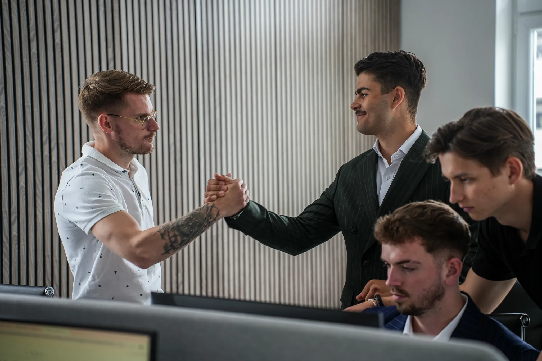 Two men shaking hands in an office while two others focus on a computer screen nearby.