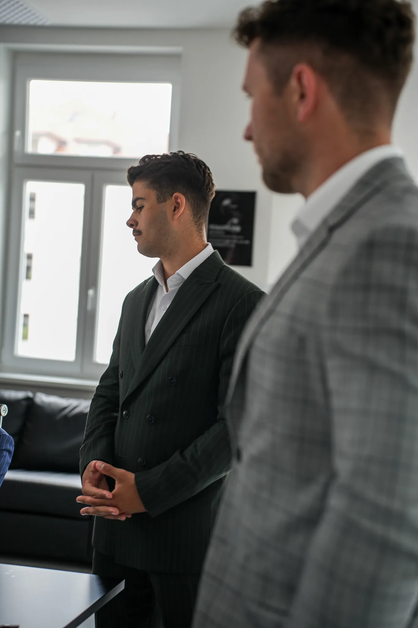 Two men in suits standing indoors near a window, one in focus wearing a dark pinstripe suit.