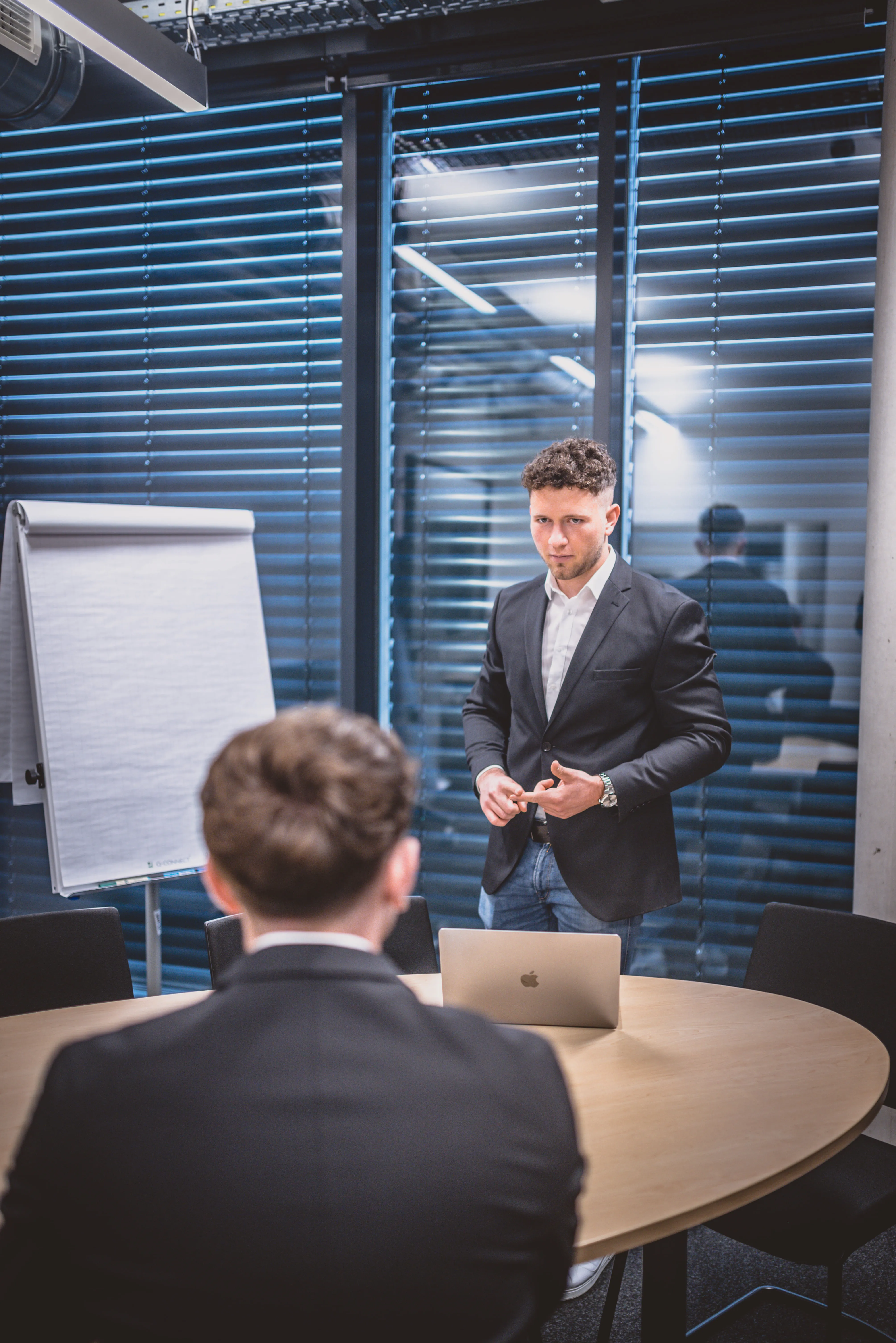 Man in suit standing and speaking in a modern office with laptop and flip chart behind him.