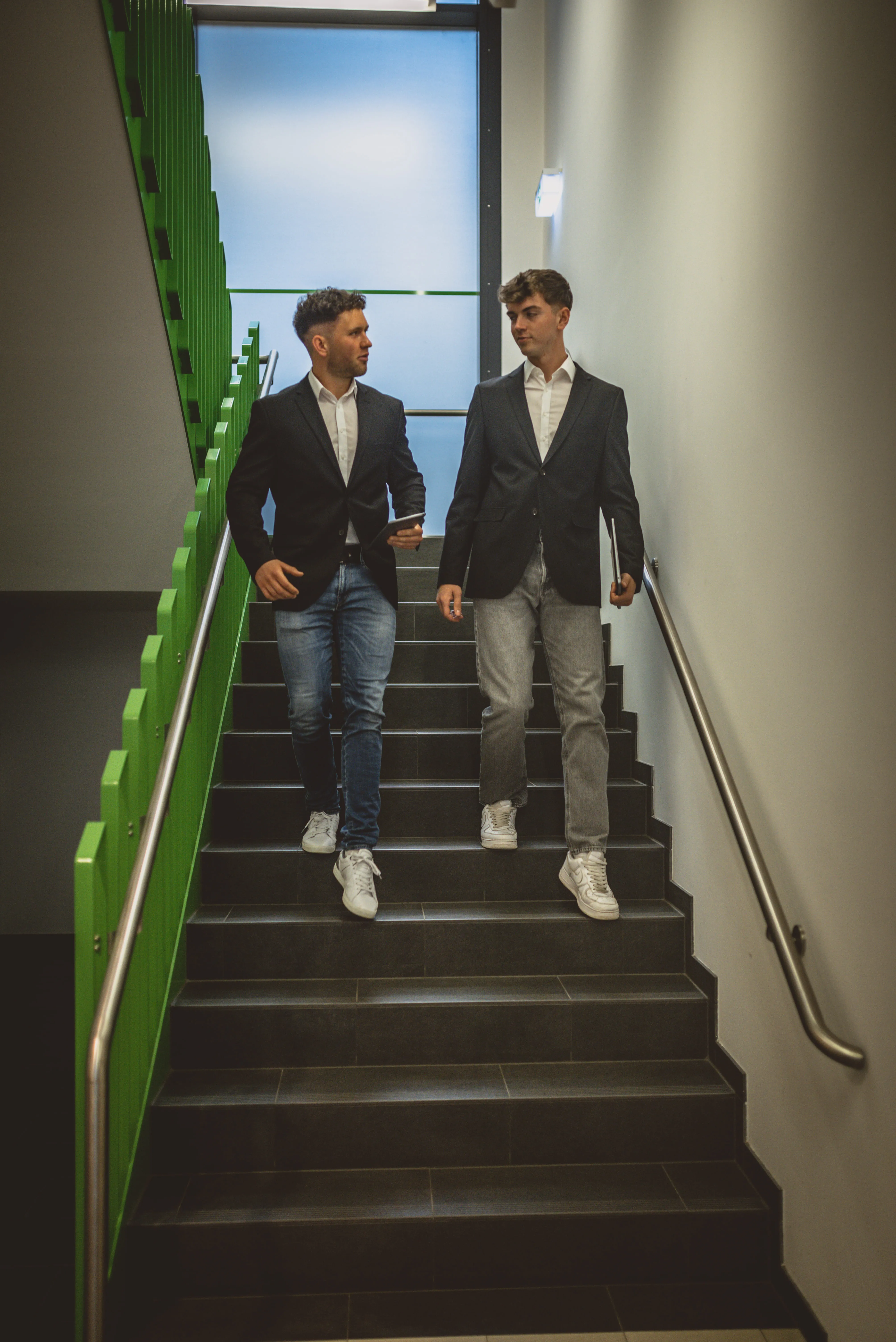 Two men in blazers and jeans walk down stairs while talking, holding tablet and laptop.