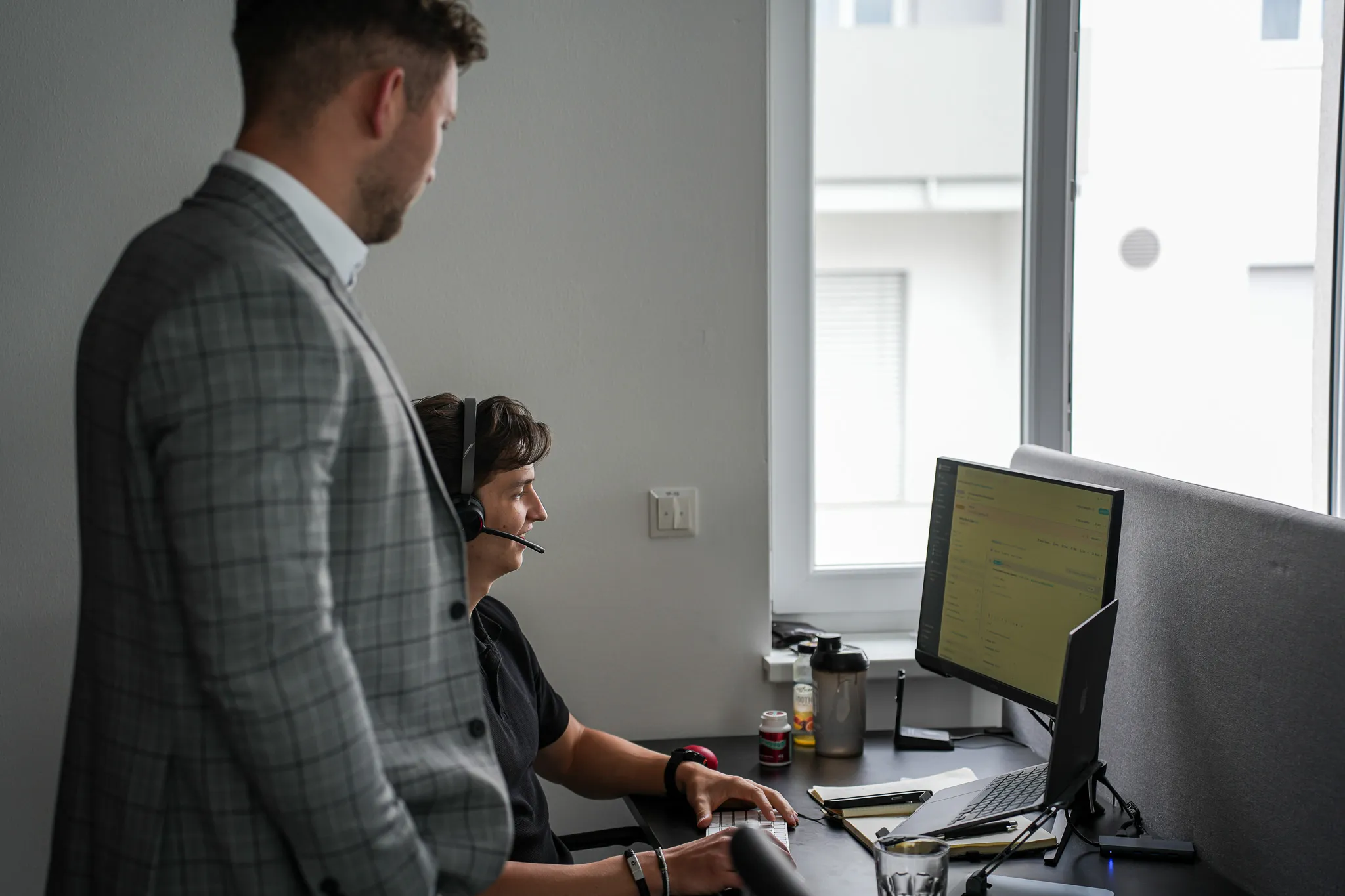 Man in headset typing on keyboard at desk while another man in plaid jacket watches.