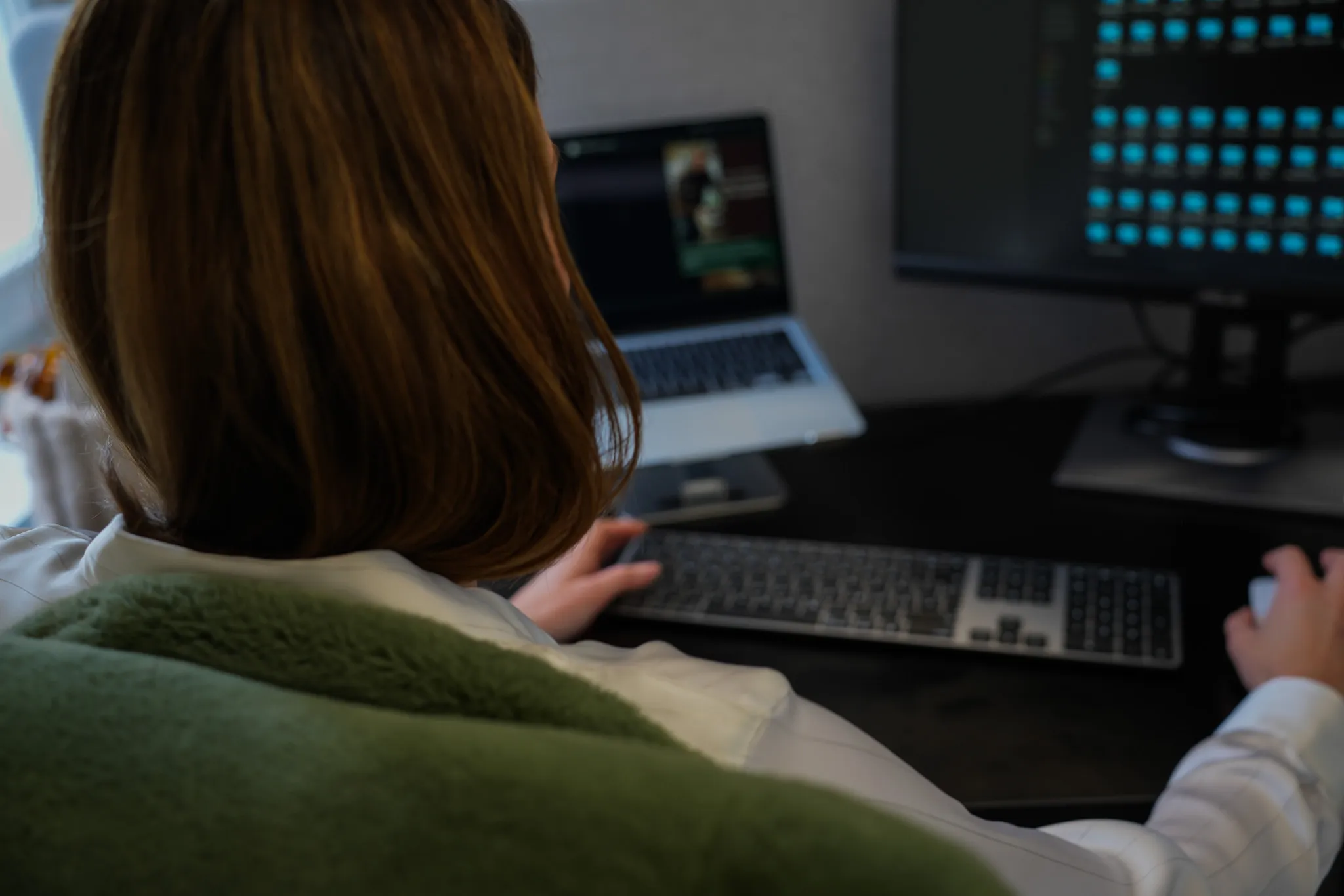 Person sitting at desk working on computer with keyboard, mouse, laptop, and monitor visible.