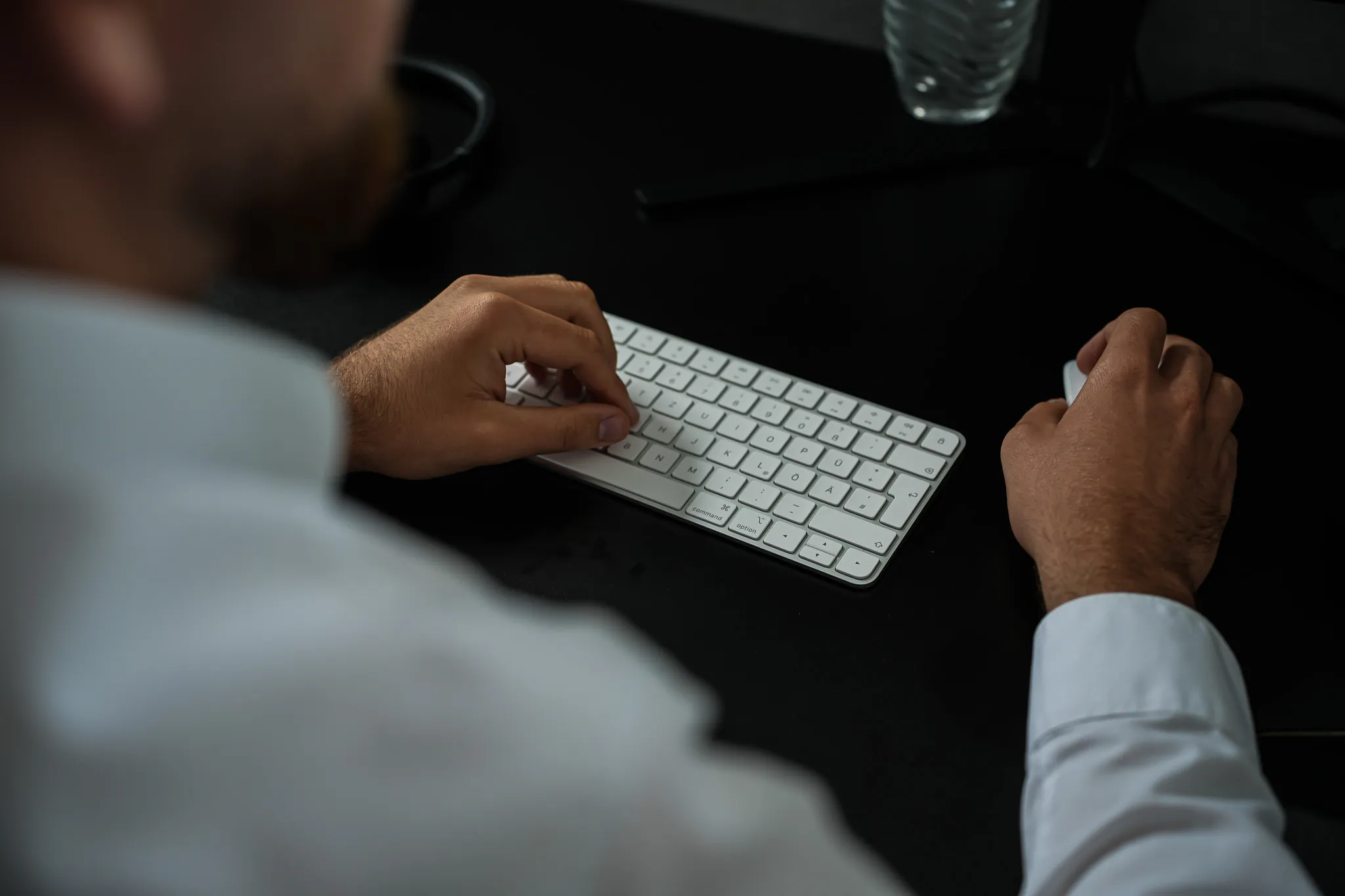 Man using a white keyboard and mouse on a dark desk, wearing a light-colored shirt.