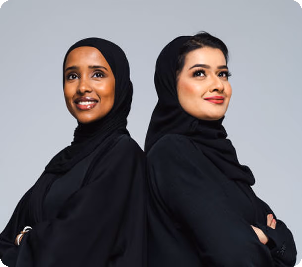 Two Muslim women in black hijabs and abayas standing back to back with arms crossed, smiling confidently.