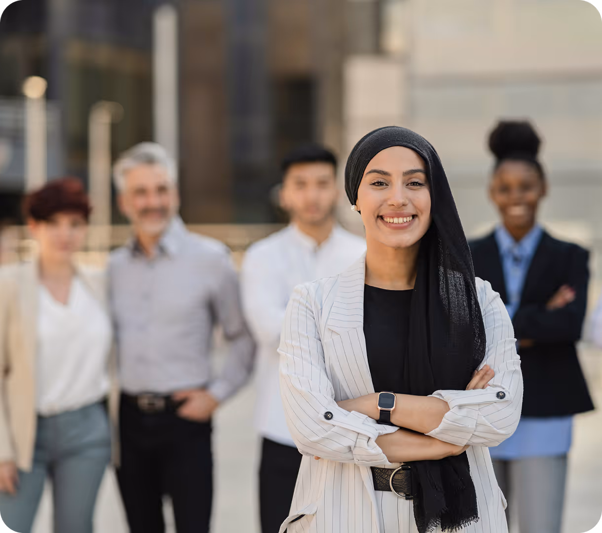 Smiling woman wearing a black hijab and white striped blazer stands confidently with arms crossed, with a diverse group of four people blurred in the background.