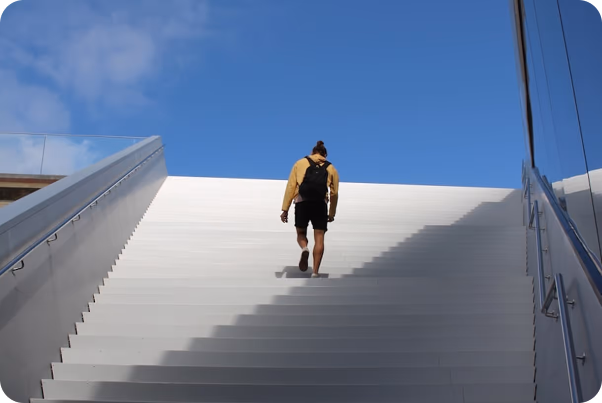 Person wearing a yellow jacket and black backpack walking up wide white stairs under a blue sky.